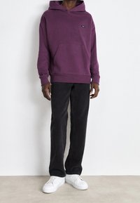 Paarse katoen hoodie met een voorzak en een klein logo, gecombineerd met zwarte spijkerbroek en witte sneakers. Eenvoudige, casual look.