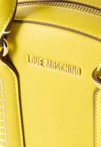 Κίτρινη τσάντα χειρός από τεχνητό δέρμα με χρυσές μεταλλικές λεπτομέρειες. Διαθέτει ανάγλυφο λογότυπο "LOVE MOSCHINO" και αντίθετη ραφή κατά μήκος των άκρων.