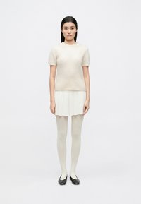 Pulover tricotat bej deschis cu mâneci scurte, asortat cu o fustă mini plisată albă, ciorapi transparenți și pantofi negri comozi. Design minimalist.