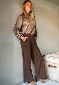 Bruine wijdvallende broeken gecombineerd met een gladde, gebroken witte blouse met een patroon en pofmouwen. Zachte texturen en neutrale tinten zijn duidelijk aanwezig.