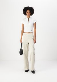 Top corto bianco a coste con maniche corte e zip frontale, abbinato a pantaloni cargo beige, borsa nera e scarpe nere.