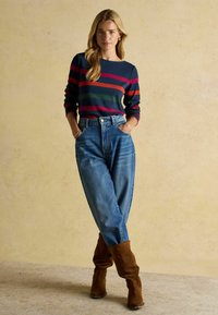 Námornícky sveter s červenými, zelenými a námorníckymi pruhmi, skombinovaný s vysokými modrými džínsami a hnedými semišovými čižmami po kolená.
