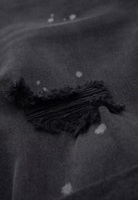 Tela negra con un desgarro deshilachado que revela los hilos subyacentes; manchas blancas dispersas realzan la textura. Material suave y de punto.