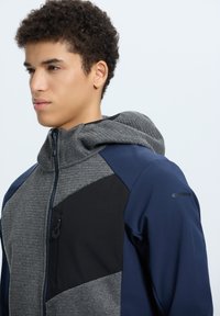 Giacca con cappuccio grigia e blu navy caratterizzata da un tessuto testurizzato, una tasca con zip e un design a blocchi di colore angolari.