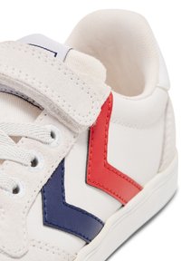 Białe sneakersy z teksturowanym zamszowym paskiem, czerwonymi i niebieskimi skórzanymi akcentami, białymi sznurowadłami i gumową podeszwą. Zbliżenie na górną część.