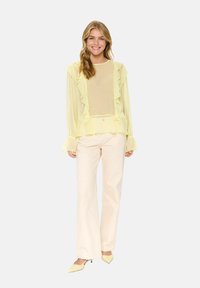 Yellow sheer blouse with ruffle details, long sleeves, and a round neckline, paired with light beige pants and yellow heels.