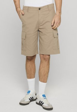 Beige cargoshorts med lommer foran og på siden, båret med hvide sokker og hvide sneakers med sorte striber og blåt logo.