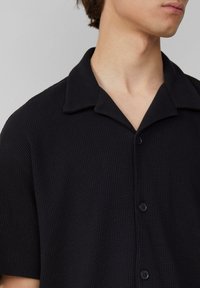 Chemise noire �à manches courtes en tissu texturé, dotée d'un col et d'une fermeture à boutons, avec un subtil motif en quadrillage.
