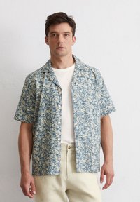Kortemouwshirt in lichtblauw met een bladmotief, voorzien van een comfortabele pasvorm, knoopsluiting en lichte stof. Gecombineerd met lichte broek.