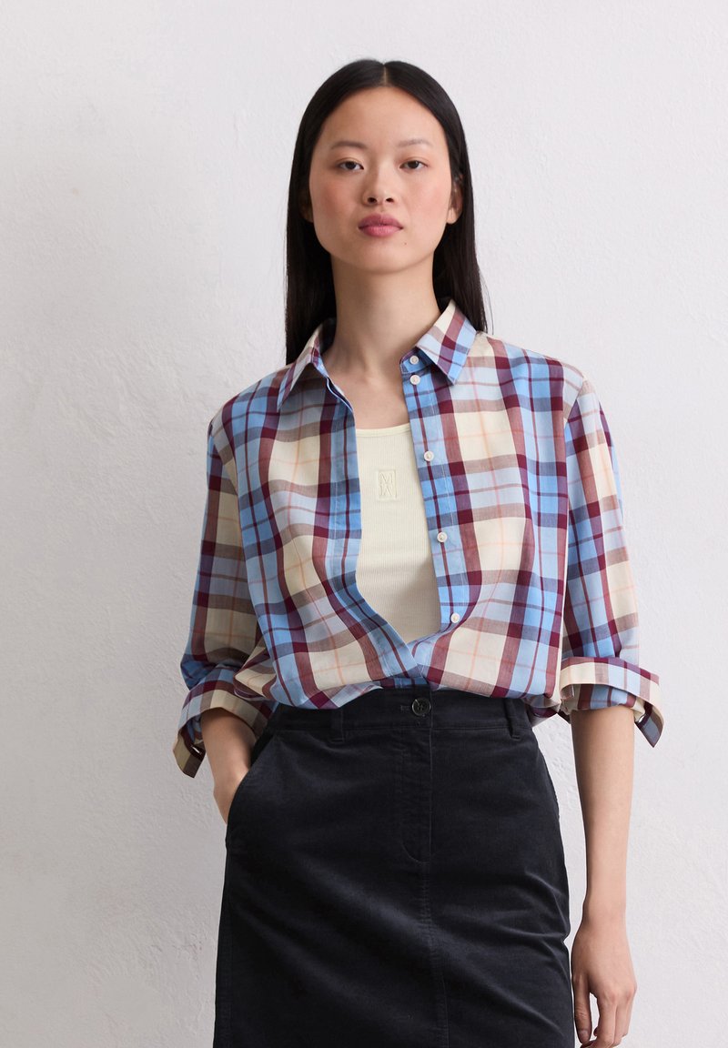 Geruit shirt in blauwe, rode en beige tinten; heeft een button-down kraag en opgerolde mouwen. Draagt over een licht tanktop en zwarte rok.