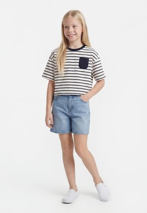 Niña sonriente con camiseta de manga corta a rayas con bolsillo azul marino, pantalones cortos vaqueros y zapatos blancos sin cordones, de pie con una mano en el bolsillo.