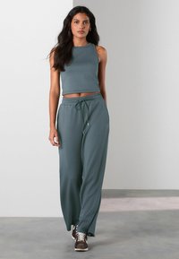 Twee-delig outfit in teal: mouwloze crop top en hoge taille broek met trekkoord. Zachte textuur; de broek heeft een relaxte pasvorm en zijzakken.