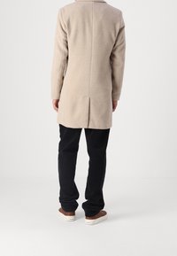Beige wool coat with long sleeves, back vent, and straight cut, paired with black pants and brown sneakers. Back view displayed.