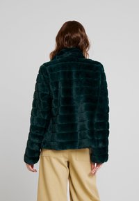 Bunda z umělé kožešiny v tmavě zelené barvě, s texturovaným pruhovaným vzorem, vysokým límcem a volným střihem. Kombinována s béžovými kalhotami.