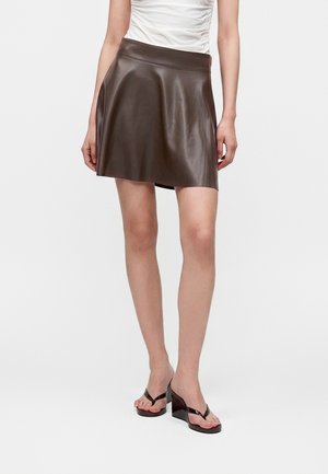 Femme portant une mini-jupe en cuir marron coupe A, un top blanc froncé et des sandales compensées noires, debout sur un fond blanc uni.