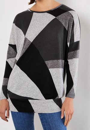 Personne portant un haut à manches longues avec de larges motifs géométriques noirs et gris et un jean bleu, vue des épaules aux hanches.