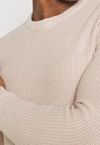 Sweter dzianinowy, w jasnym beżowym kolorze, z prążkowanym okrągłym dekoltem i teksturowanym wzorem, z zbliżeniem na detal rękawa.
