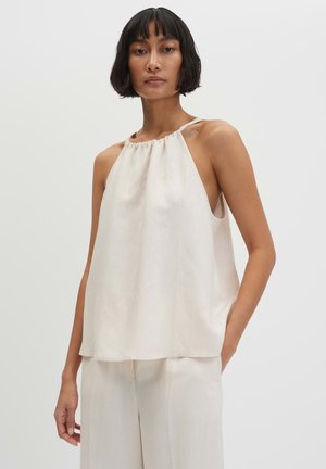 Donna con top halter crema senza maniche e pantaloni crema a gamba larga abbinati, in piedi con una mano in tasca.