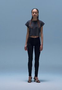 Juoda trumpa palaidinė, juodos aukšto liemens siauros džinsai ir juodi atviri bateliai su aukštu kulnu. Modelis turi ilgus pintus plaukus ir stovi prie šviesiai mėlyno fono.