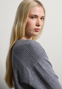 Grijze gebreide trui met geribbelde schouderdetails. Zachte textuur, ontspannen pasvorm en een gladde afwerking, gedragen door een model met lang haar.