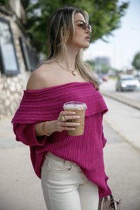 Túlméretezett, bordázott magenta pulóver vállra omló nyakkivágással. Világos színű farmernadrággal viseli, kezében egy átlátszó pohár jéghideg kávéval.