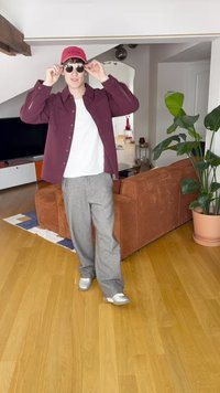 Tumši sarkans pogājams krekls virs baltas T-krekla, pelēkas rūtainas bikses, sudraba krāsas kedas, sarkana cepure un apaļas saulesbrilles, stāvot viesistabā.