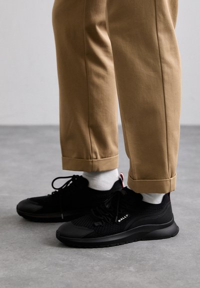 Black mesh and synthetic athletic shoes with textured upper, round toe, and flat rubber sole. Paired with light brown cuffed trousers.