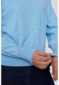 Gaiši zils adīts džemperis ar teksturētu mazāku caurumu rakstu. Ribotas aproces un apakšmala. Gluda auduma ar brīvu piegriezumu.