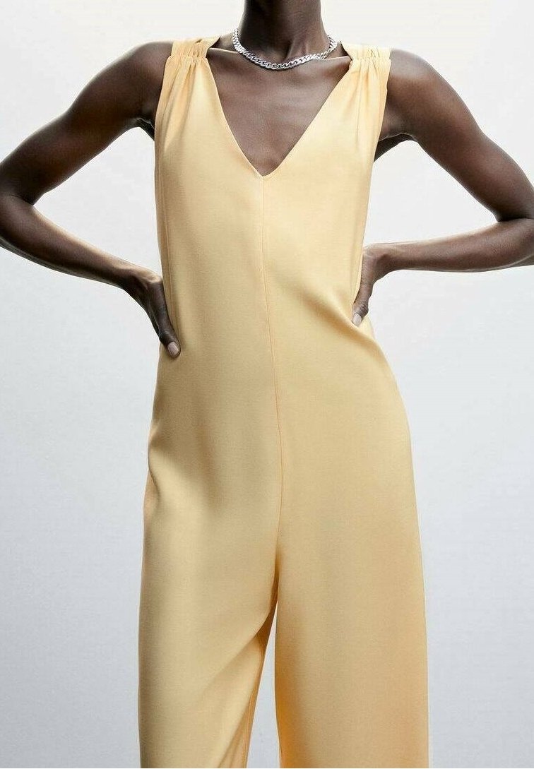 Personne mince portant une combinaison jaune clair sans manches avec un col en V profond et un collier chaîne argenté, mains sur les hanches sur un fond uni.