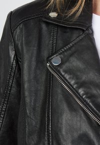 Svart läderjacka med en texturerad yta, diagonal dragkedja och reflekterande silverdetaljer på kragen och axeln.