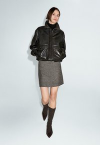 Giacca di pelle nera con due tasche frontali, indossata sopra una gonna grigia aderente. La modella indossa calze scure fino al ginocchio e scarpe con la punta. Sfondo semplice.
