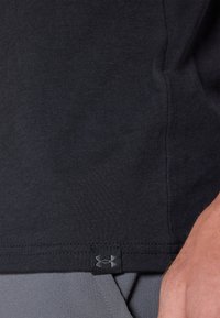 Fekete, rövid ujjú, puha anyagból készült póló, melynek alján egy kis Under Armour logó található, szürke sportnadrággal párosítva.