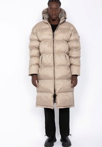 Palton puffer bej, lung, cu secțiuni quilted orizontale, glugă, fermoar frontal și buzunare laterale, purtat peste o ținută neagră și botine.