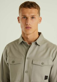 Šviesiai pilka marškinėliai su užsegimu, pagaminti iš sklandžios medžiagos, turi dvi krūtinės kišenes ir logotipo pleistrą apatinėje dešinėje pusėje. Klasikinio tipo apykaklė.