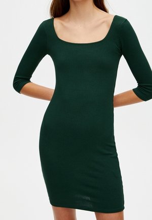 Žena oblečená v přiléhavých žebrovaných tmavě zelených šatech se tříčtvrtečními rukávy a širokým čtvercovým výstřihem, stojící s rukama za zády.