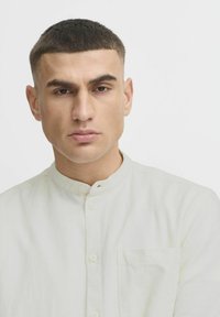 Camisa de lino color beige claro con cuello alzado, parte delantera abotonada y un bolsillo en el pecho. Textura suave con un ligero brillo.