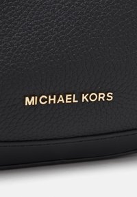 Mustat nahkalaukku, jossa on teksturoitu pinta, kultainen "MICHAEL KORS" -brändäys ja sileä alareuna.