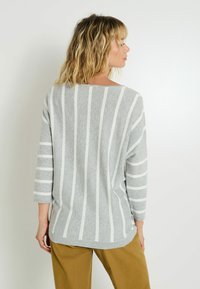 Mujer con cabello rubio ondulado hasta los hombros, vistiendo un suéter a rayas gris y blanco y pantalones marrones, de espaldas frente a una pared lisa.