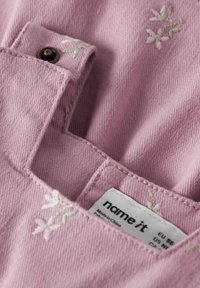 Lyserødt stof i close-up med synlig tekstur, med blomsterbroderi og en etiket syet på siden.