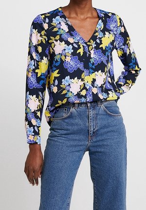 Kvinde iført en marineblå blomstret langærmet bluse med gule, blå og lyserøde blomster kombineret med højtaljede blå jeans.