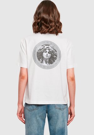 Vit bomull t-shirt med en cirkulär grå grafik av en kvinna med flödande hår på baksidan, inramad med ett grekiskt nyckelmönster.