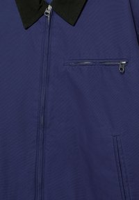 Granatowa kurtka z czarnym kołnierzem, zamkiem błyskawicznym z przodu, zapinaną na zamek kieszenią na piersi oraz pionową kieszenią boczną.