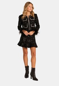 Zwarte paillettenjurk met een aansluitend ontwerp, ritssluiting aan de voorkant, veren manchet en een gerimpelde zoom. Gepaard met zwarte enkellaarsjes.