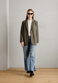 Ελαιοπράσινο oversized σακάκι, λευκό μπλουζάκι, ανοιχτό μπλε φαρδιά τζιν και μαύρα παπούτσια. Μοντέλο που φορά μαύρα γυαλιά ηλίου και στέκεται σε ξύλινο δάπεδο.