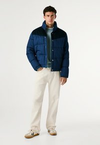 Modra puhovka z temno modrim delom na rami, s pritiskači in teksturirano tkanino, kombinirana s smetanastimi hlačami in športnimi copati z vzorcem.