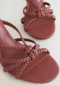 Geflochtene burgunderfarbene Sandalen mit flacher Sohle, die über aufwendige geflochtene Riemen und eine geknotete Akzentuierung an der Vorderseite verfügen. Strukturierte Lederoberfläche.