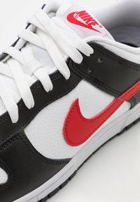 Basket Nike blanche, noire et rouge avec des lacets blancs, un logo swoosh rouge sur le côté et un marquage Nike rouge sur la languette.