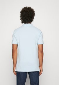 Ljusblå bomullspolo med korta ärmar, krage och rak nederkant, i kombination med mörka jeans. Bakifrån utan synliga logotyper.