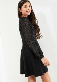 Robe noire avec un corsage ajusté et une jupe évasée. Les manches longues présentent un tissu texturé et froncé. Tissu de jupe lisse et uni.