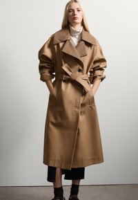 Abrigo trench marrón con un gran cuello de cuadros, mangas anchas, un cinturón atado y mínimos botones. Lleva puesto sobre unos jeans oscuros y zapatos marrones.